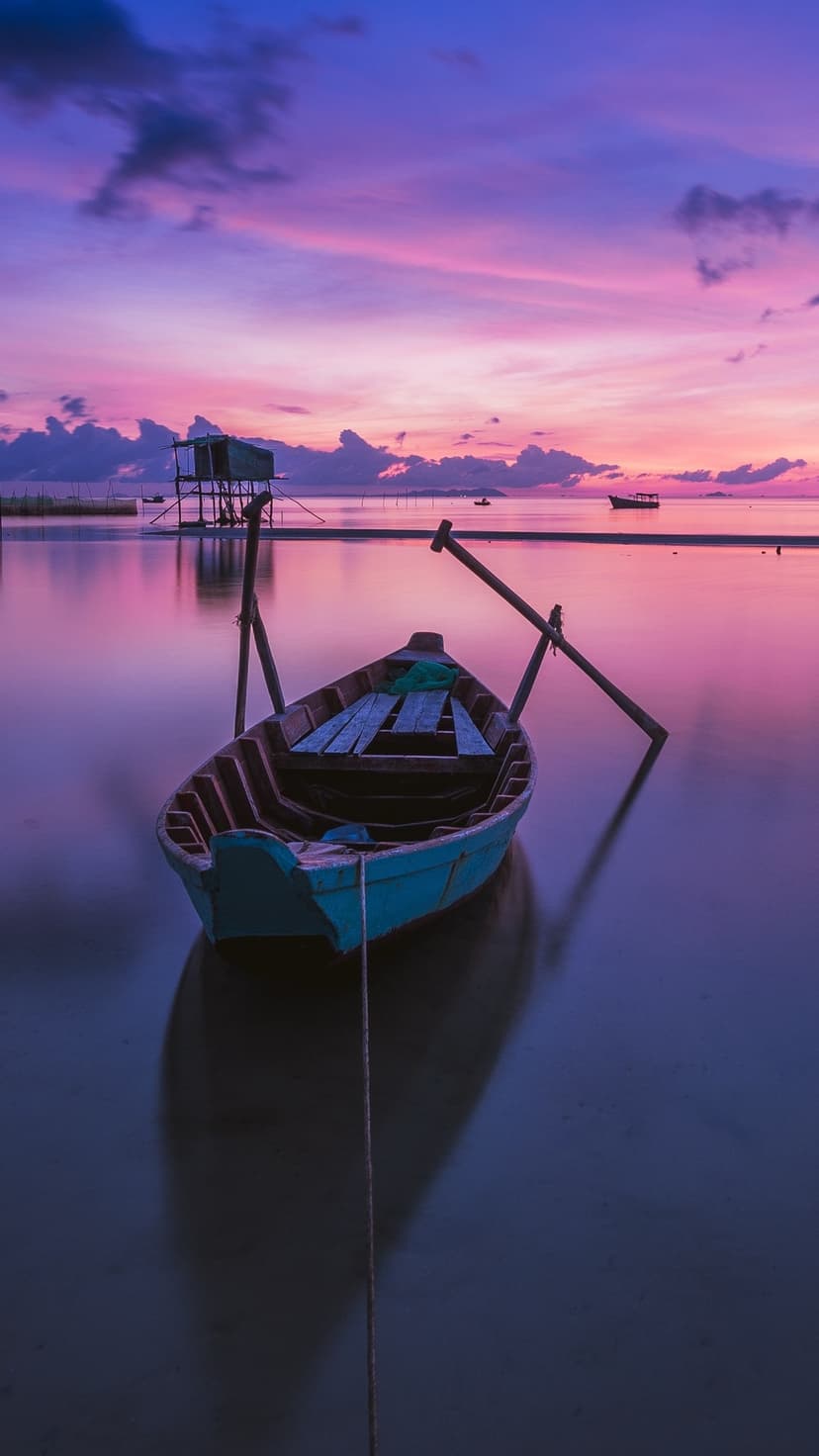 Teal wooden boat on calm water under a vibrant purple and pink sunset sky with distant stilt house silhouettes.