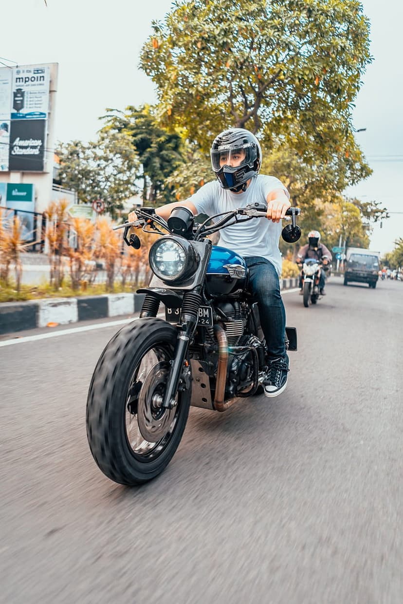 Man in a helmet rides a custom motorcycle with a large front tire on a paved city street with motion blur.