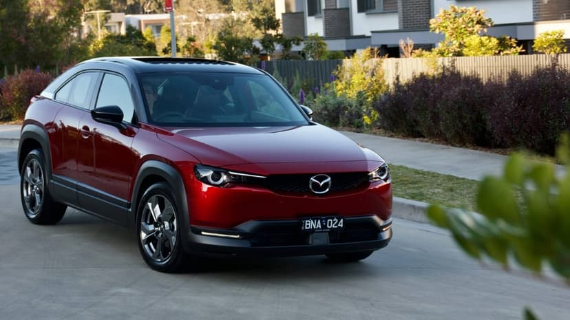 Red Mazda MX-30 SUV driving on a suburban road with sunlight reflecting off its metallic paint and black roof.