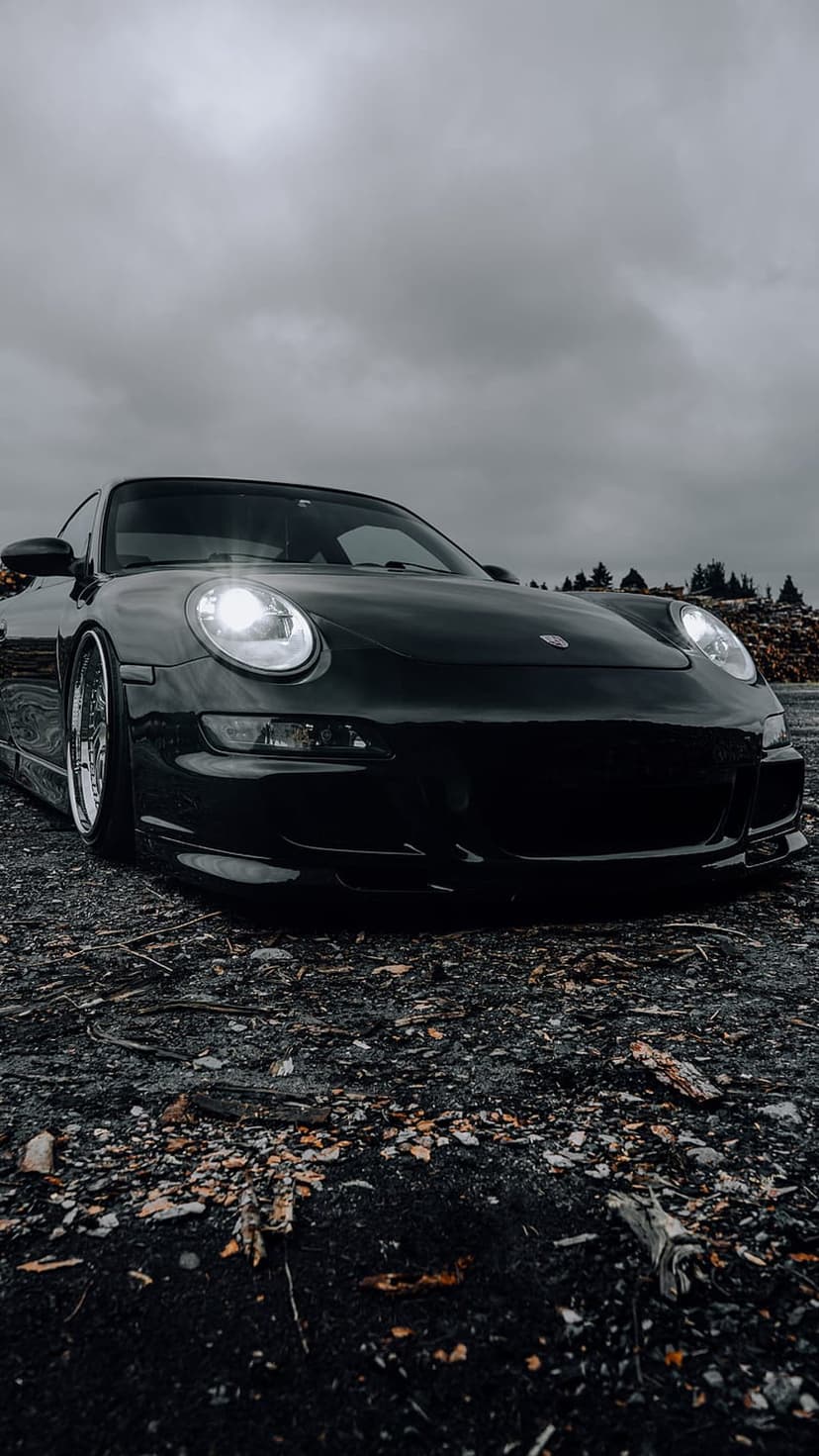 Black Porsche 911 with glowing headlights on a gravel road under a dark overcast sky in a low angle shot.