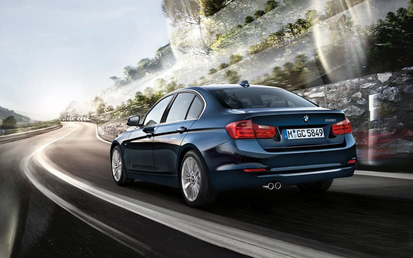 Dark blue BMW 328i luxury sedan driving on a winding mountain road with motion blur and bright sun flares.