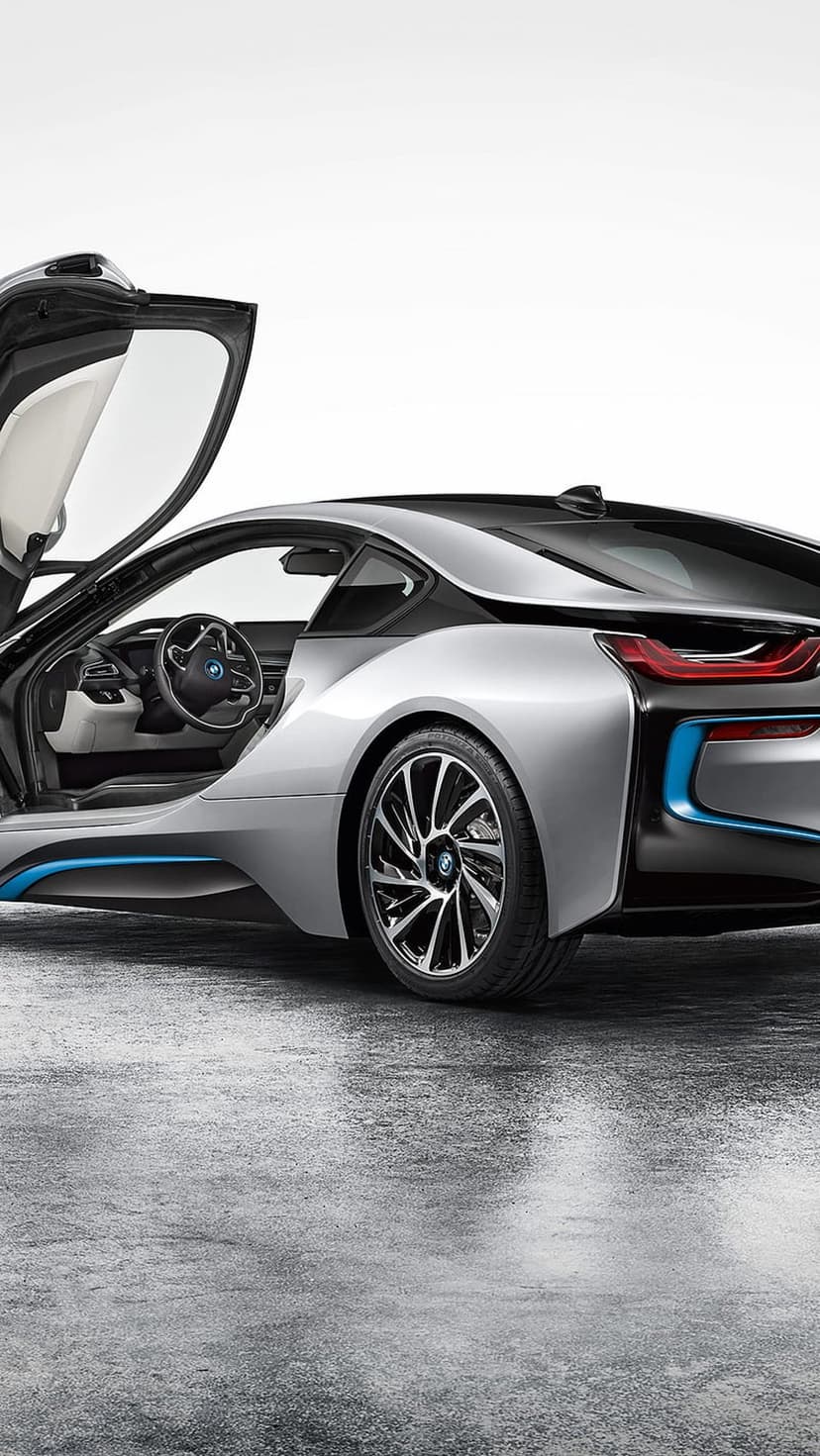 Silver BMW i8 sports car with open butterfly doors parked on wet pavement reflecting blue accent lights.