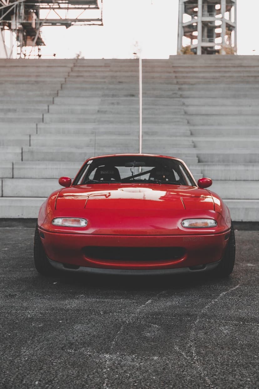 Red Mazda Miata NA front profile parked in front of grey concrete stadium steps under soft daylight.