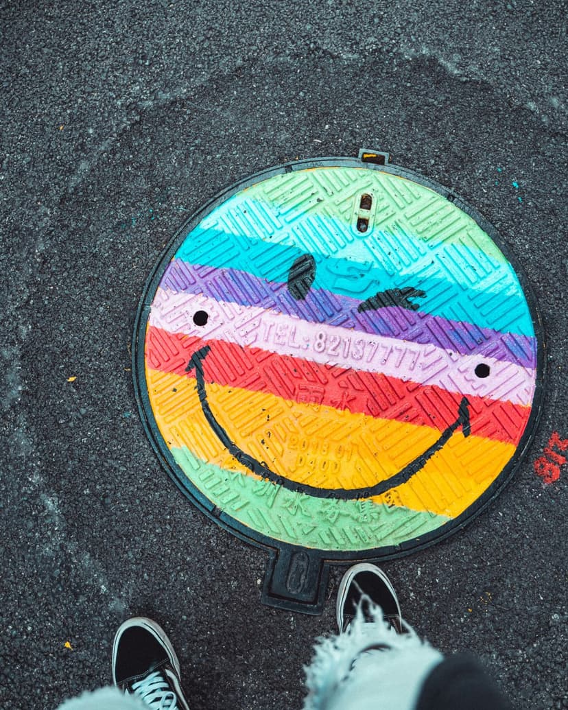 Smiley face manhole cover with rainbow stripes on dark asphalt next to two pairs of sneakers in a city street.