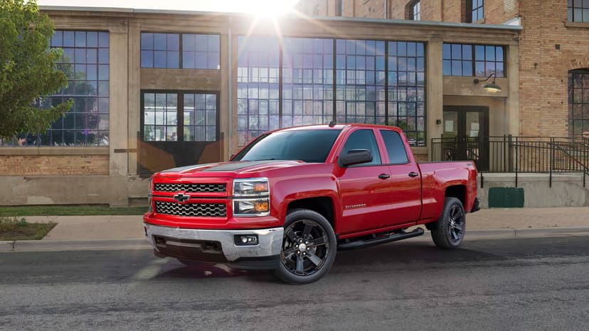 Red Chevrolet Silverado parked on a street with black hood stripes, chrome bumpers, and sun flare background.