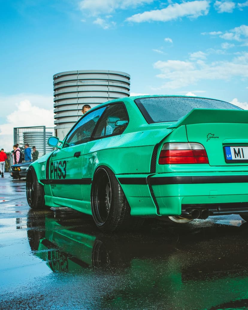 Green BMW M3 E36 sports car with a rear wing and wide fenders parked on reflective wet asphalt in daylight.