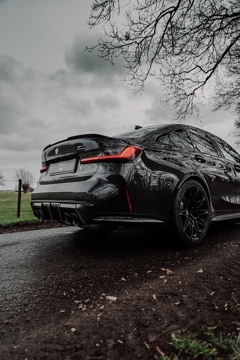 Black BMW M3 Coupe with red taillights and quad exhaust pipes on a wet road under a cloudy sky with bare trees.