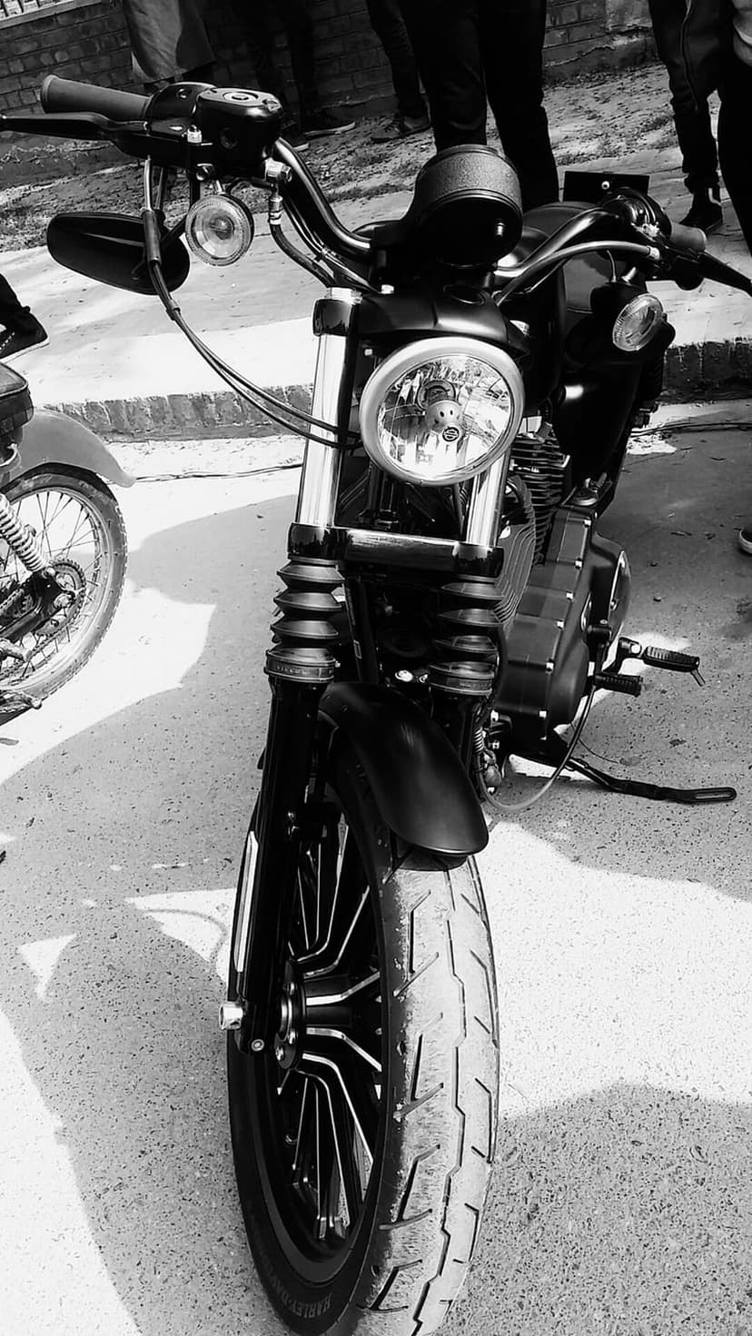 Vintage motorcycle front wheel and headlight casting long shadows on asphalt in a high-contrast monochrome shot.