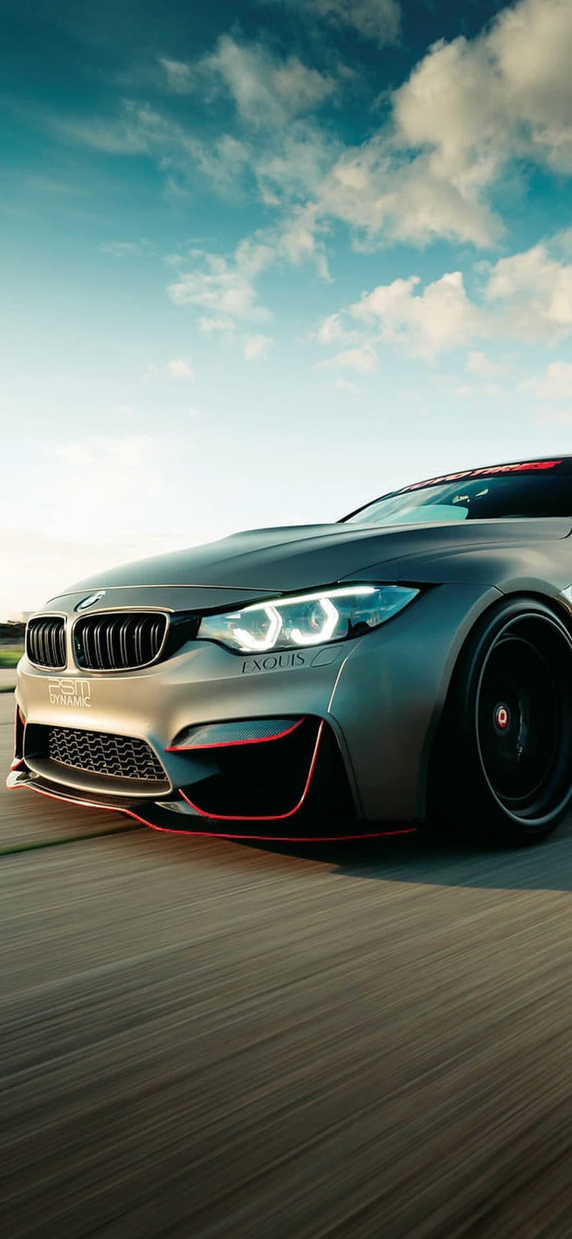 Grey BMW M4 with red accents and glowing headlights driving on a road under a moody, overcast cloudy sky.