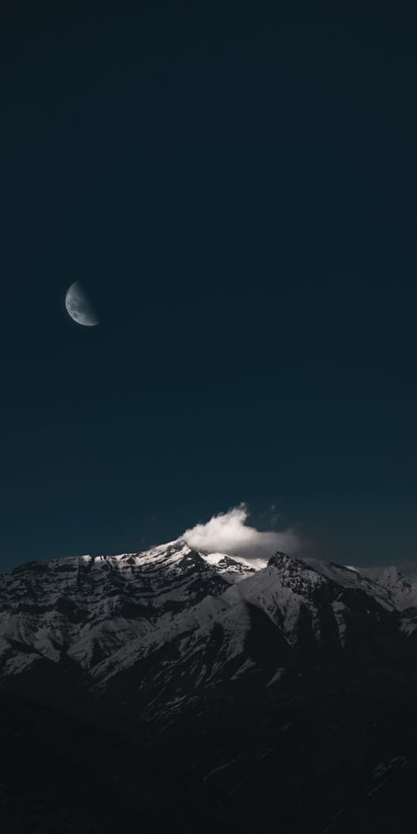 Snow-capped mountain peaks beneath a thin crescent moon and a single white cloud in a dark blue twilight sky.