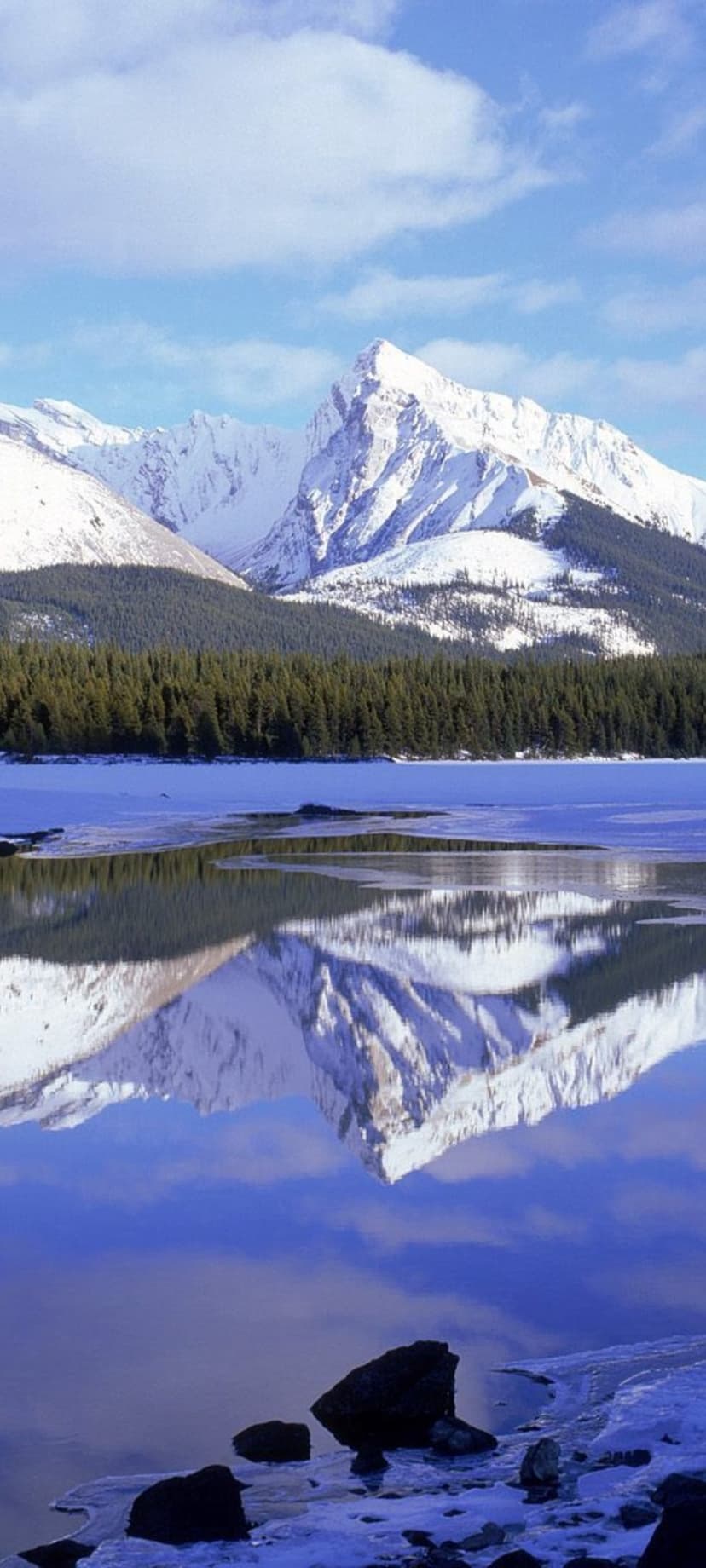 Snow-capped mountain peaks reflecting in a still blue lake surrounded by dark green pine trees under a soft sky.