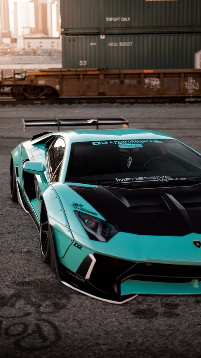 Teal Lamborghini Aventador with a widebody kit parked on asphalt near graffiti and shipping containers.