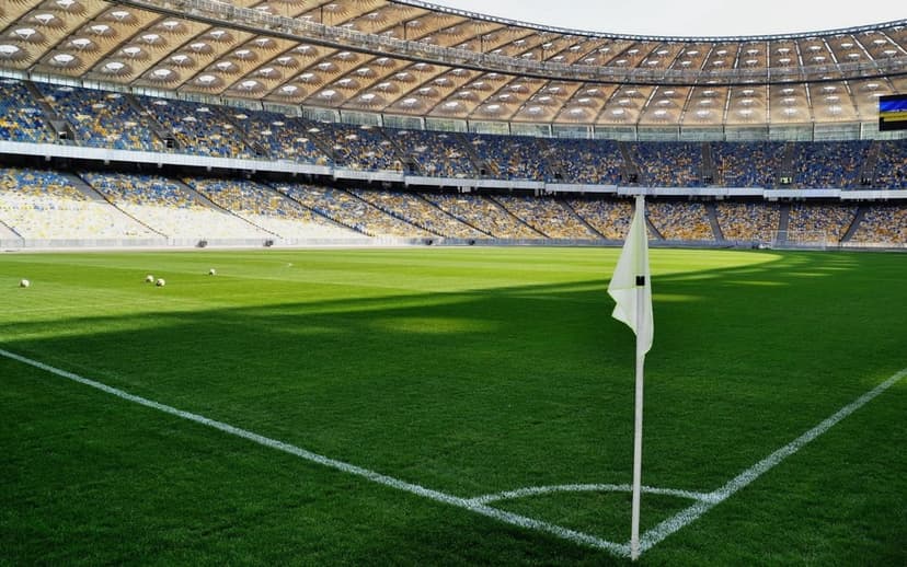 White corner flag on a green grass pitch with rows of yellow and blue stadium seating in the sunny background.