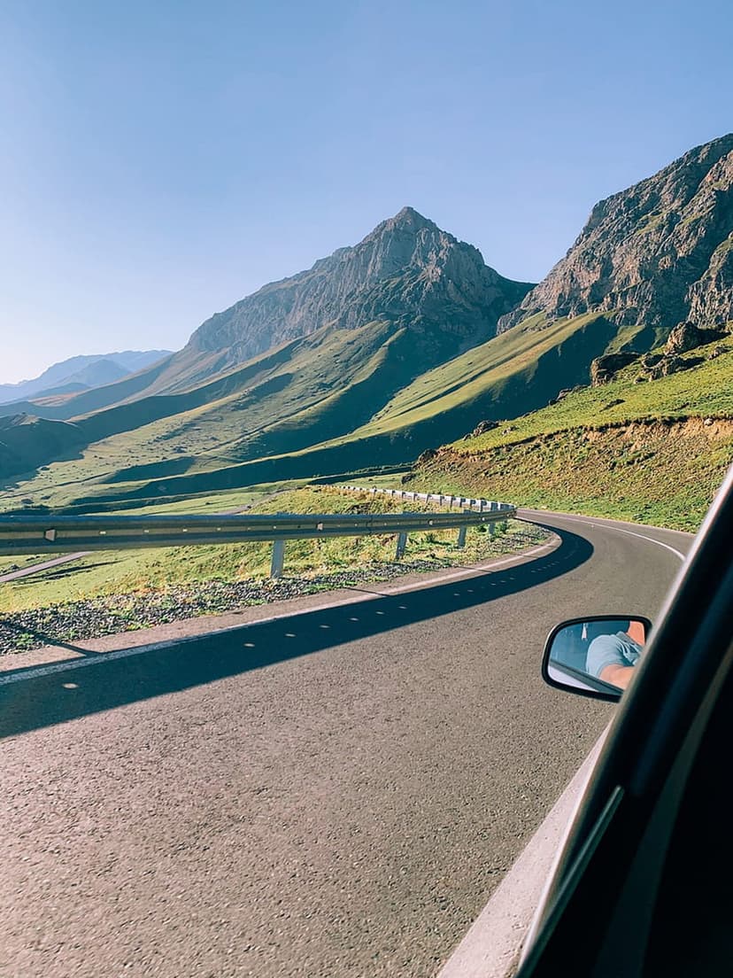 Winding asphalt road leads toward rugged mountain peaks under a clear blue sky seen from a car passenger seat.