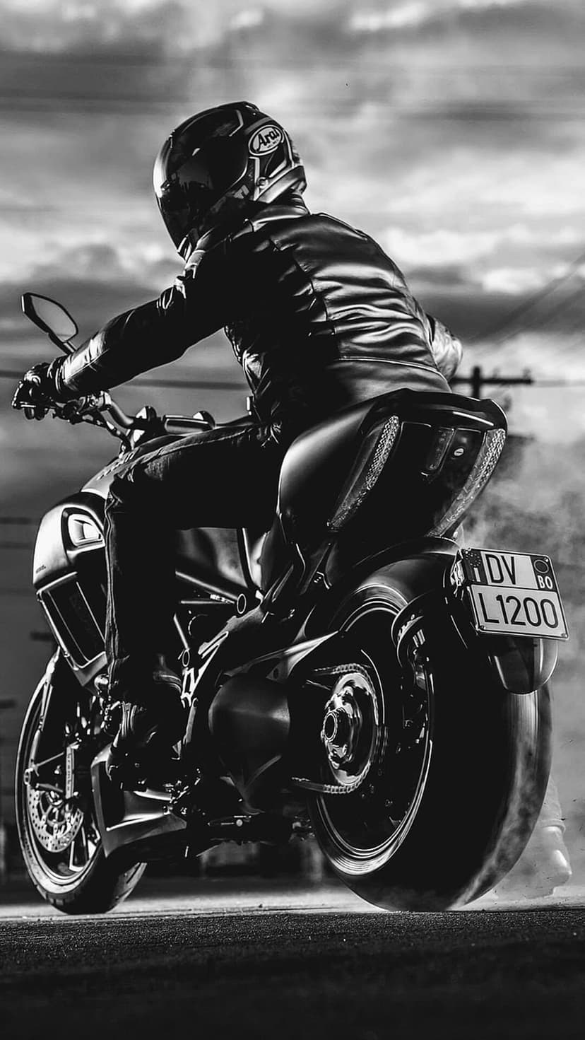 Motorcyclist in leather gear leans a sportbike into a turn as smoke rises from the tire in monochrome tones.