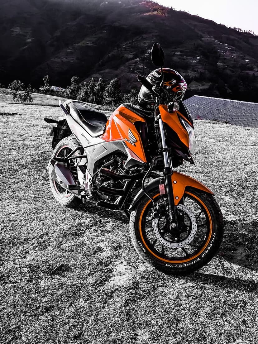 Orange motorcycle with a black helmet on a grassy hill before a desaturated mountain range under a bright sky.