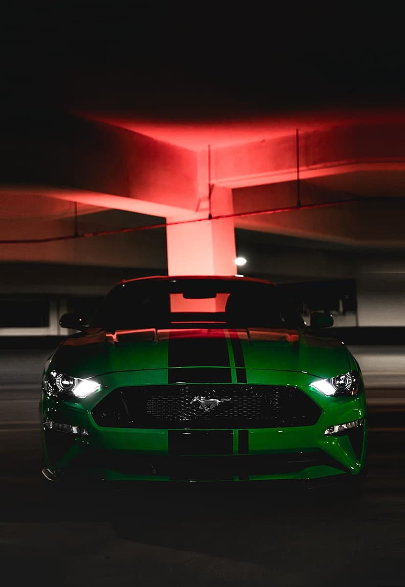Green Ford Mustang with black stripes and glowing headlights parked in a dark garage with red ambient lighting.