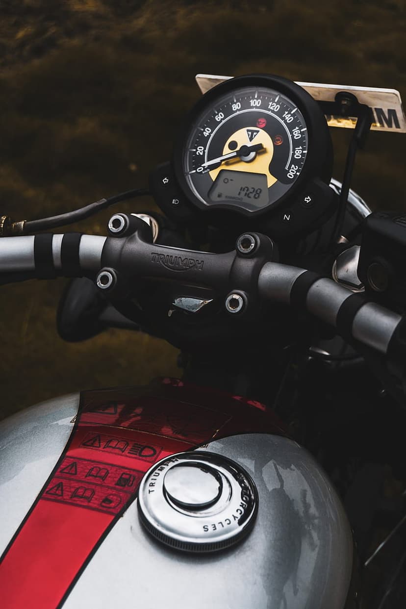 Triumph motorcycle dashboard with a chrome fuel tank, analog speedometer, and red stripe detailing on silver metal.