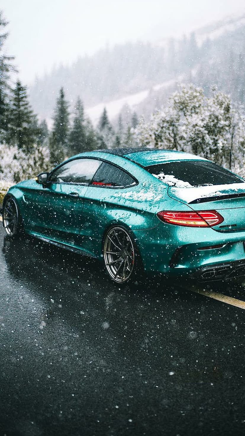 Teal sports coupe on a wet asphalt road surrounded by snow-covered trees and a misty mountain backdrop.