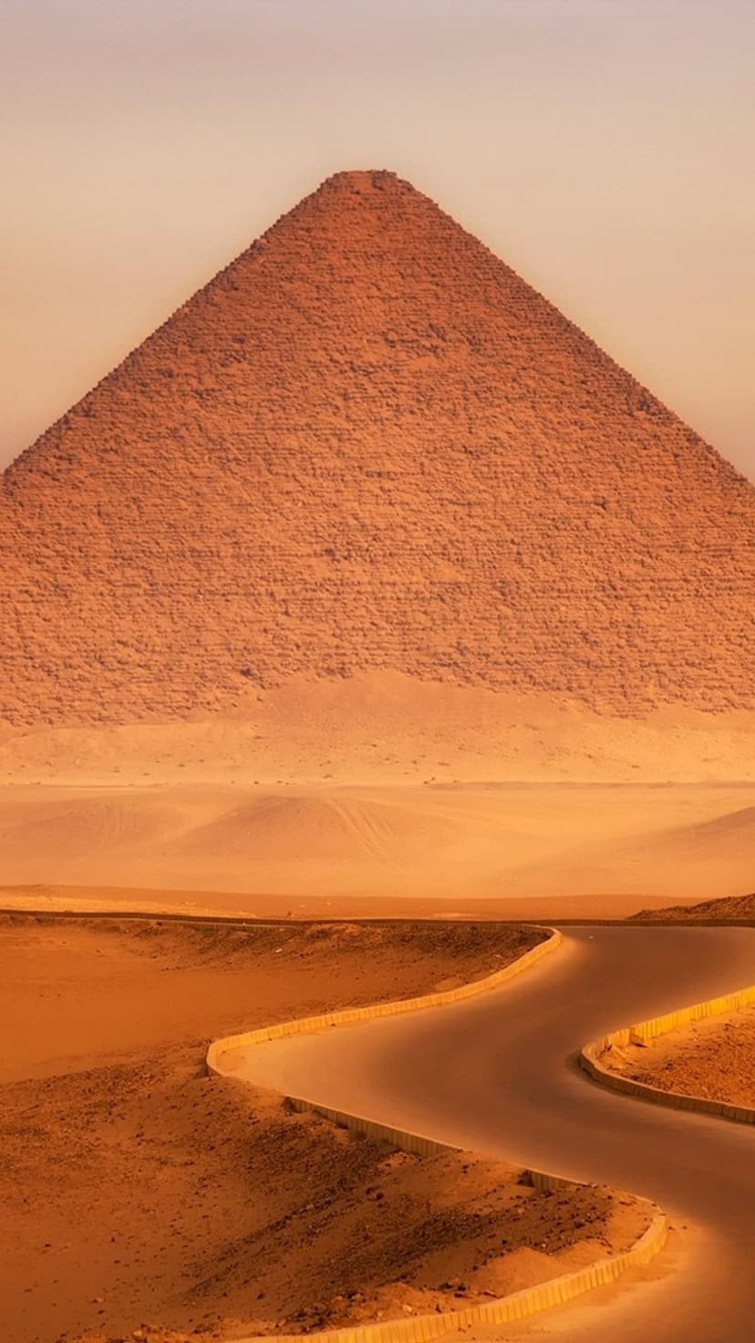 The Red Pyramid stands amidst dusty sand dunes and a winding road under a warm, hazy Egyptian desert sky.