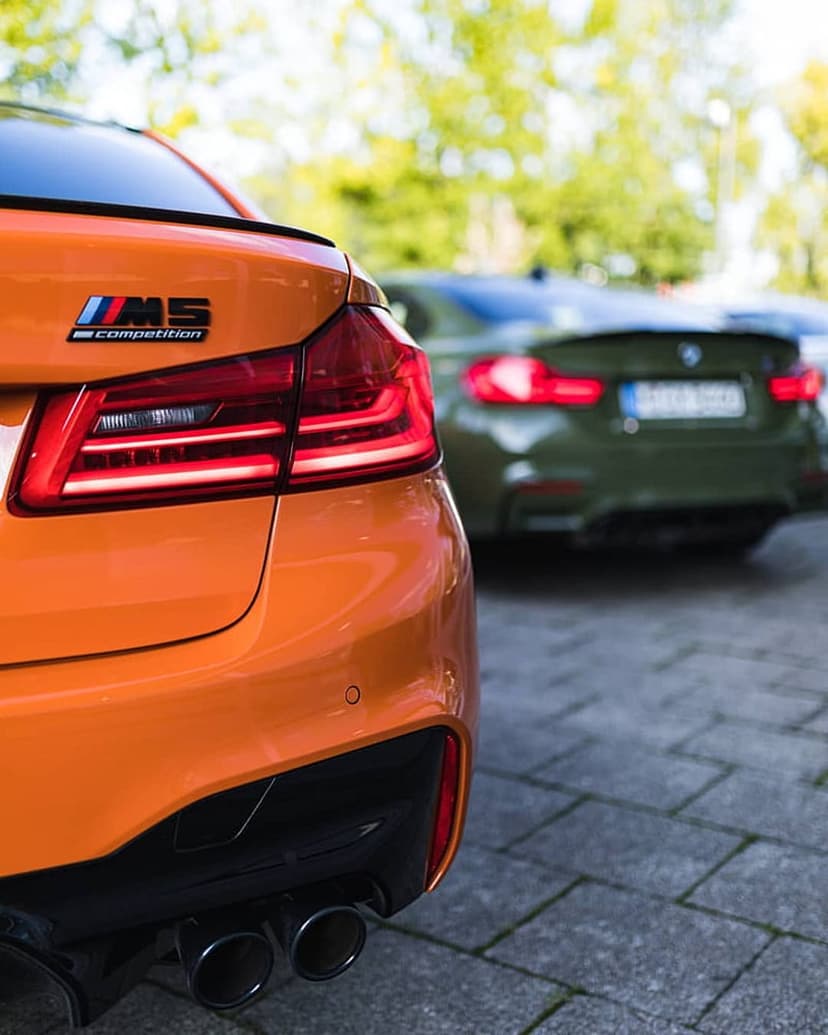 Orange BMW M5 Competition rear showing LED taillights and quad exhausts with a green car in the background.
