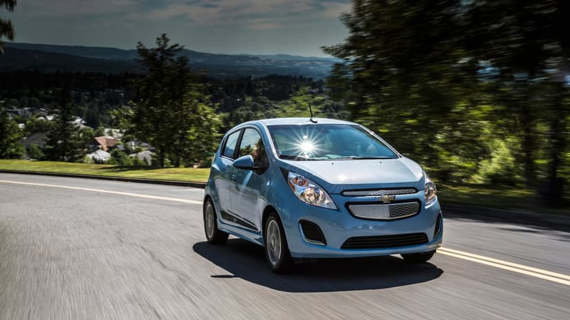 Light blue Chevrolet Spark EV driving on a winding road with a sun glare on the windshield and green trees.