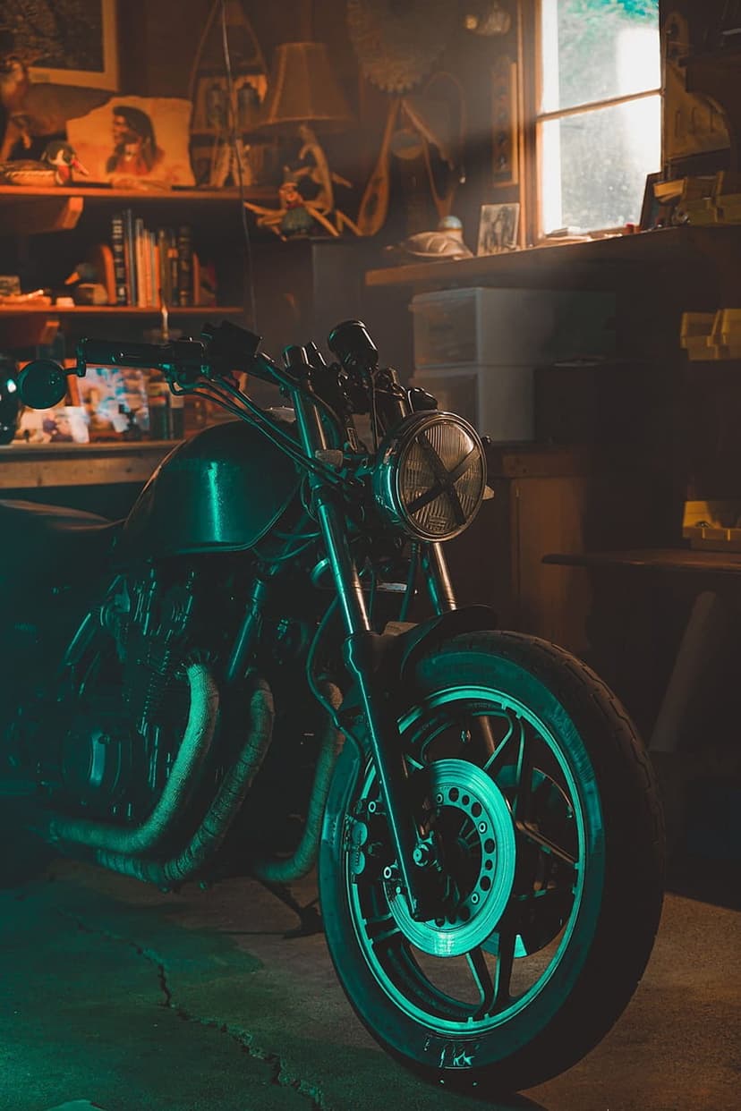 Vintage motorcycle engine and front wheel illuminated by teal light in a cluttered mechanical garage workshop.