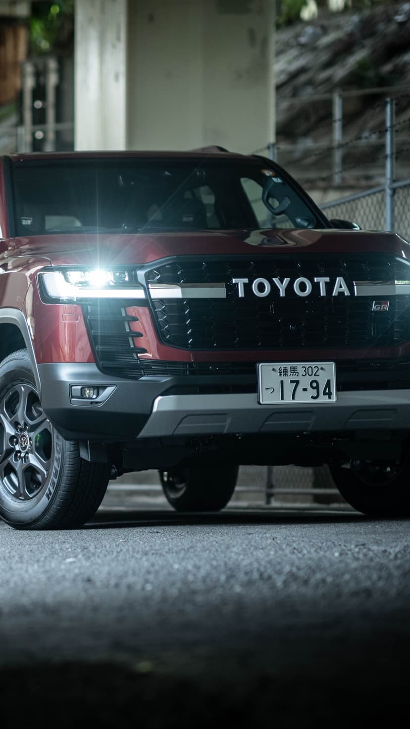 Close-up of a red Toyota Land Cruiser GR Sport with glowing headlights and black TOYOTA grille under a bridge.