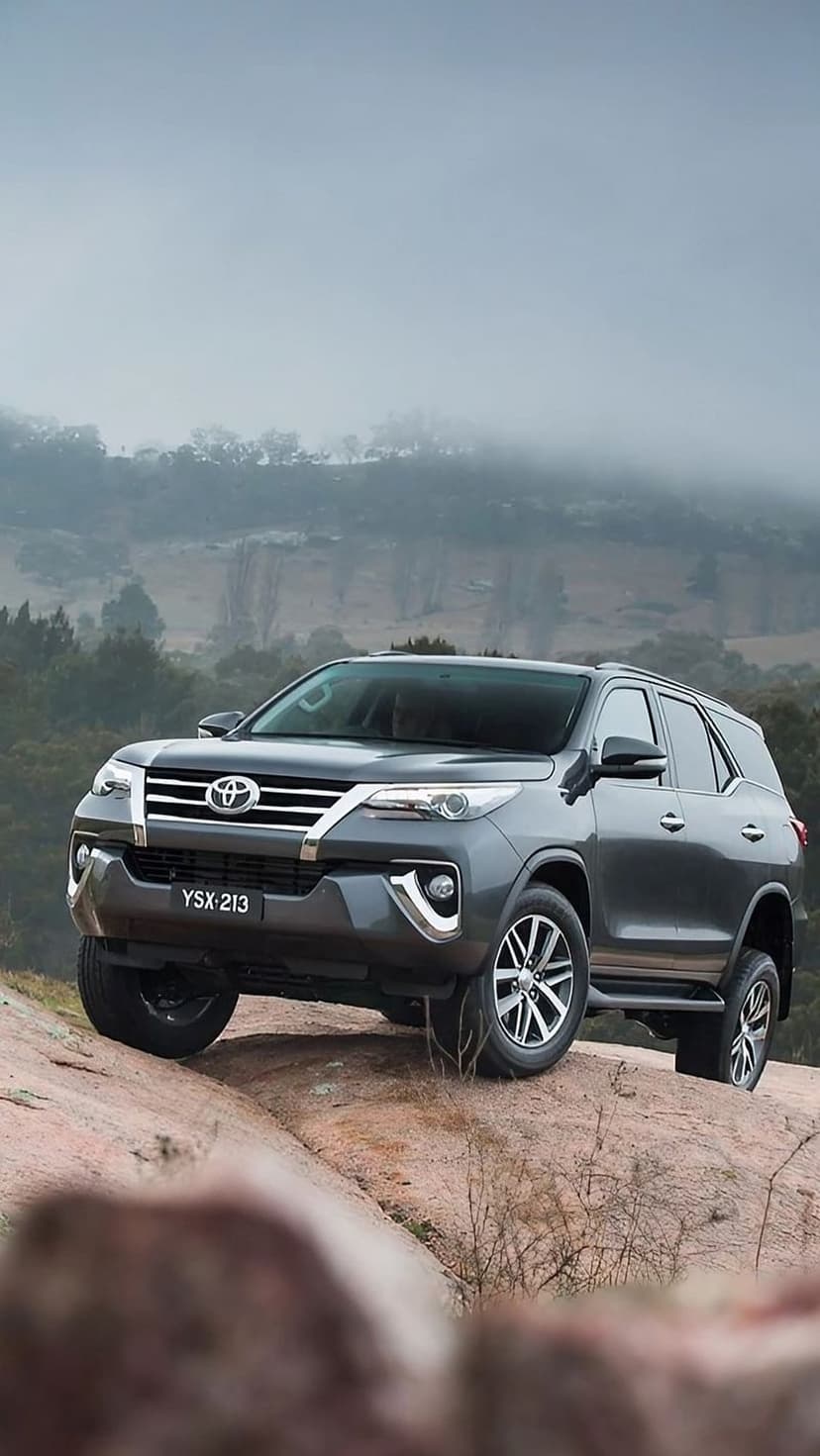 Toyota Fortuner SUV climbing a steep rocky path with misty mountains and evergreen trees in the background.