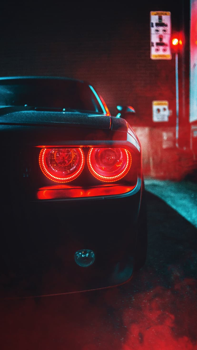 Circular red LED taillights on a dark muscle car reflecting blue neon light against a smoky brick wall at night.
