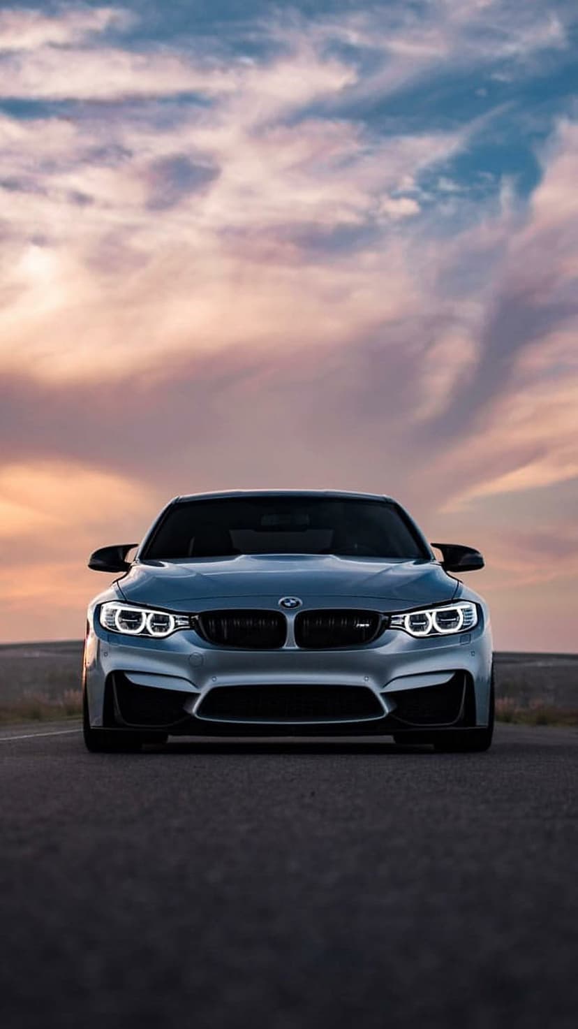 BMW M4 sports car front grille and glowing LED headlights on an asphalt road under a colorful sunset sky.