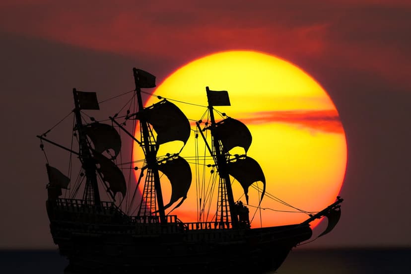 Sailing ship silhouette with intricate rigging against a massive glowing sun and a vibrant orange sunset sky.