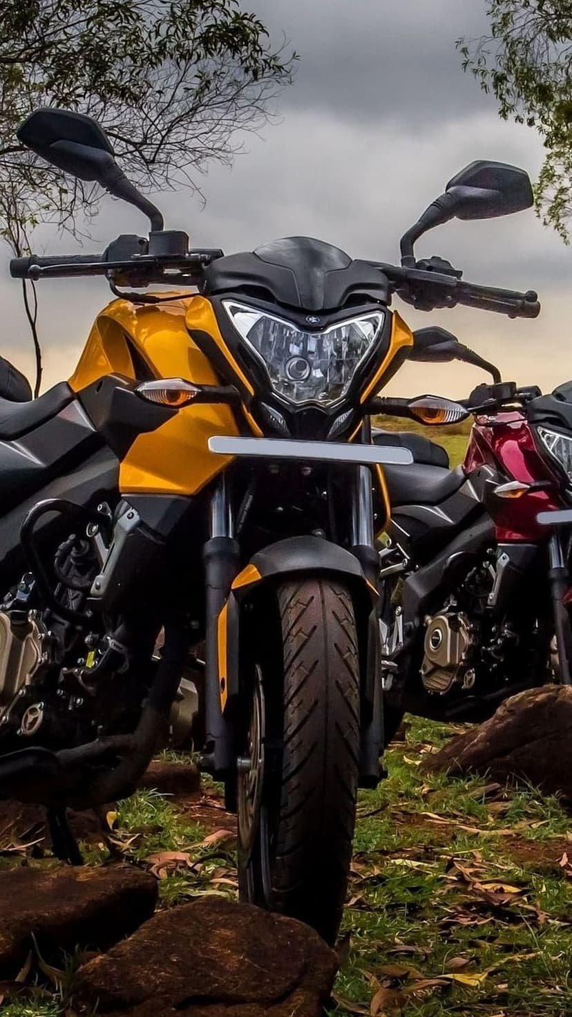 Yellow and red Bajaj Pulsar NS200 motorcycles parked on rocky green grass beneath a cloudy, overcast sky.