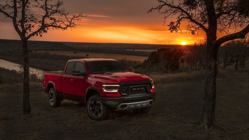 Red pickup truck parked on a hill between two silhouetted trees against a bright orange sunset over a valley.