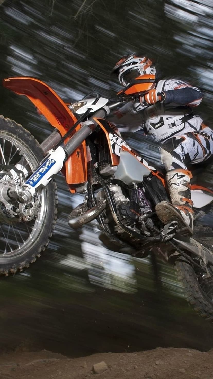 Motocross rider in orange gear jumping a dirt bike mid-air through a blurred forest on a portrait background.
