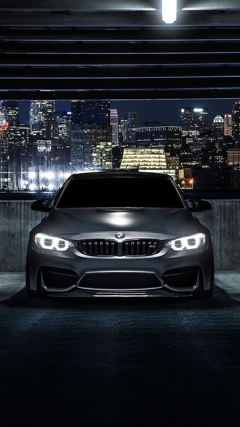 BMW M4 coupe with illuminated headlights parked on a garage roof overlooking a luminous night city skyline.