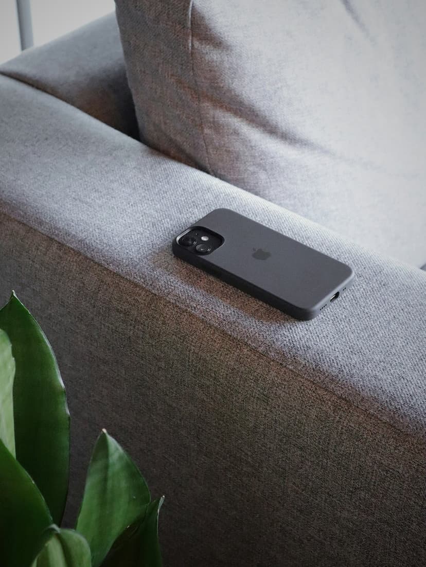 Dark gray smartphone sits on a textured charcoal sofa armrest behind out-of-focus green indoor plant leaves.