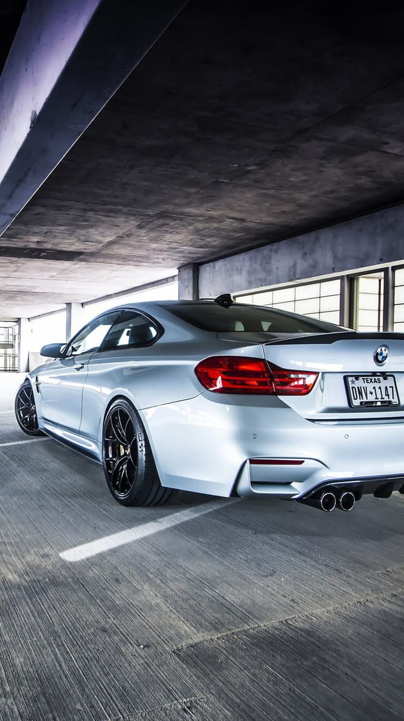 Silver BMW M4 Coupe rear view showing black rims and Texas license plate in a concrete parking garage setting.