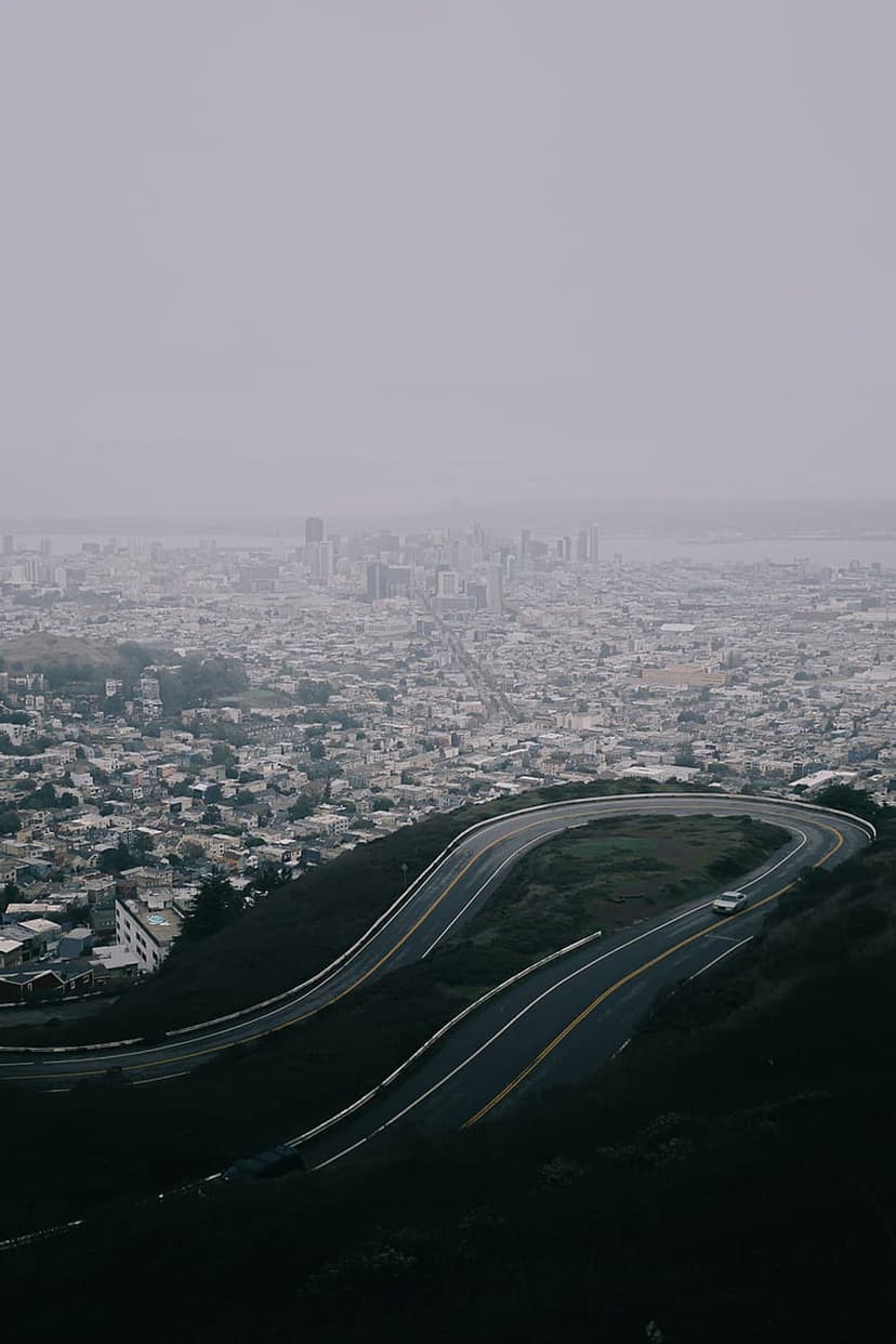 Winding mountain road curves toward a foggy San Francisco cityscape with a single car driving on the asphalt.
