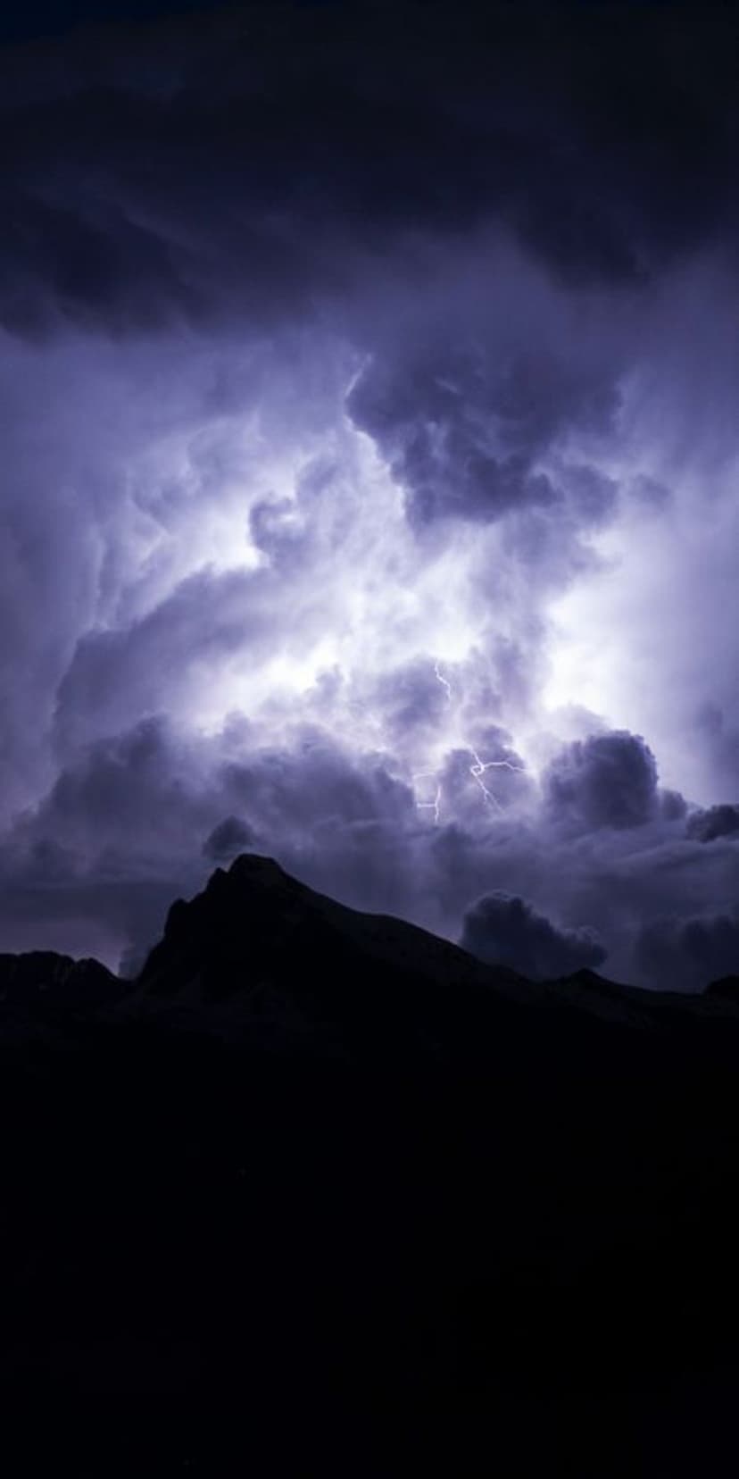 Bright lightning bolt striking through dark storm clouds above the sharp silhouettes of rugged mountain peaks.