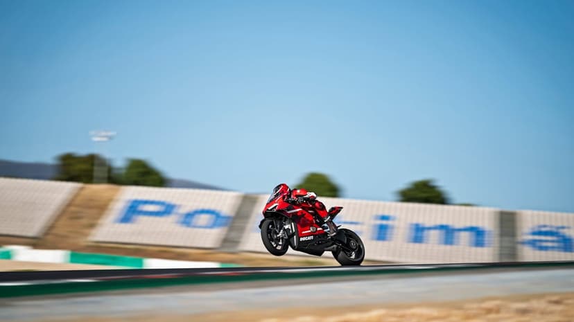 Red Ducati motorcycle jumps over a track crest with motion-blurred grandstands and blue sky background.