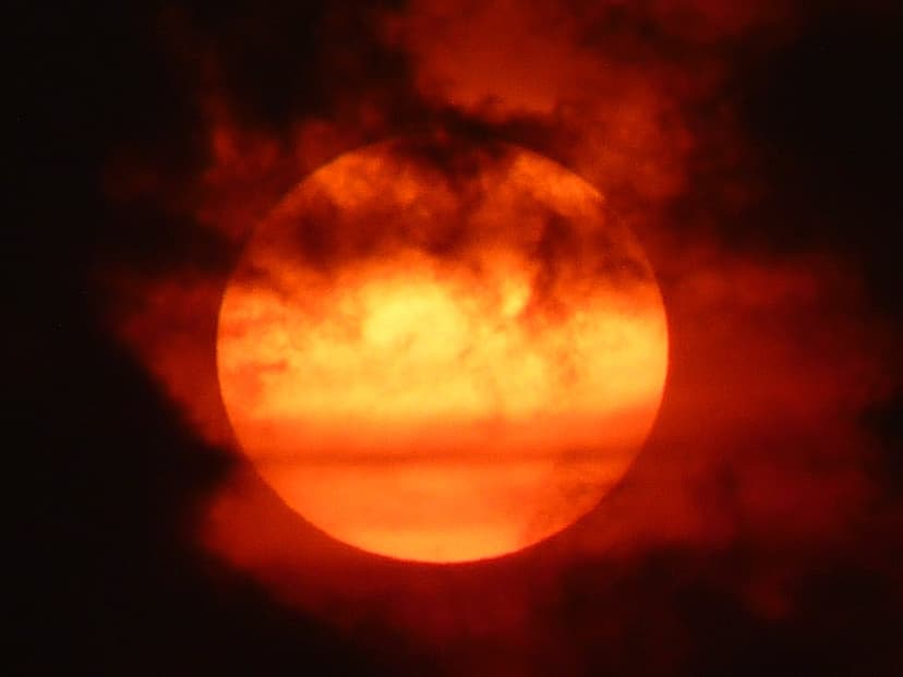 Vibrant orange sun orb glows behind dark wispy clouds during a dramatic golden hour in the evening sky.