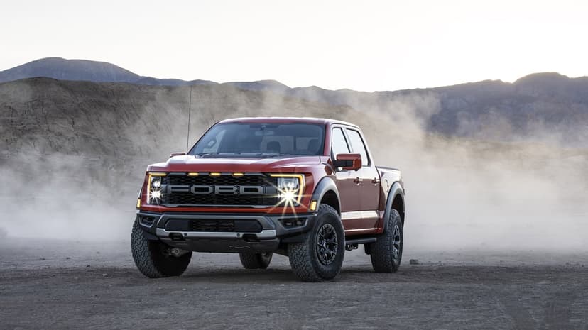 Red Ford F-150 Raptor truck in a dusty desert with headlights on against a mountain backdrop.