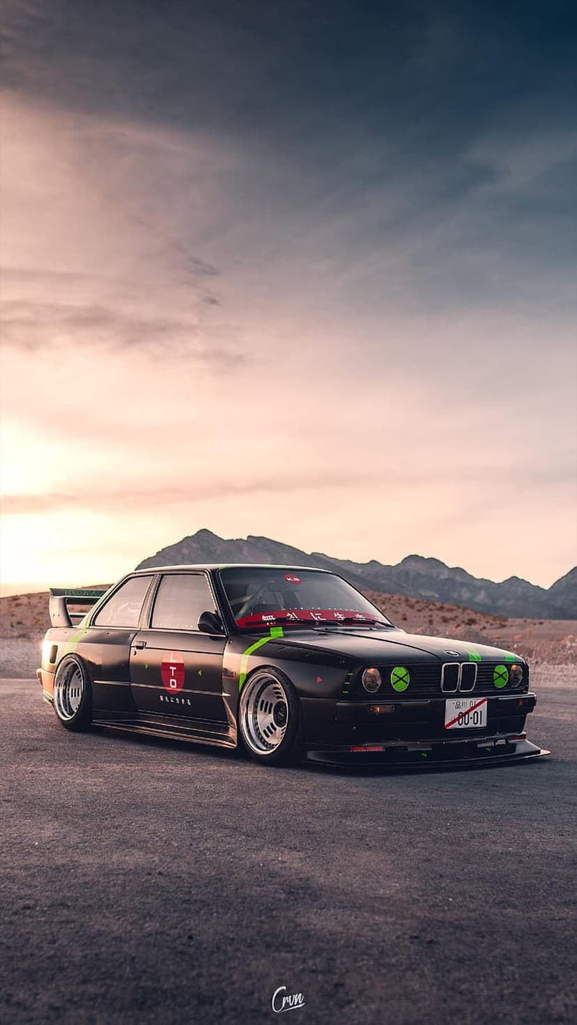 Black BMW E30 M3 car with green accents parked on a dusty desert road against a sunset mountain landscape backdrop.