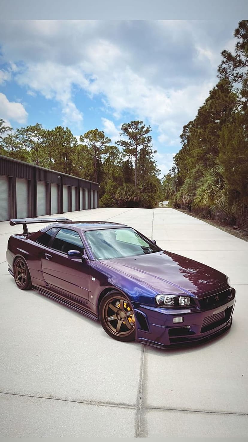 Purple Nissan Skyline R34 GT-R with a large rear wing parked on concrete in front of storage units and trees.