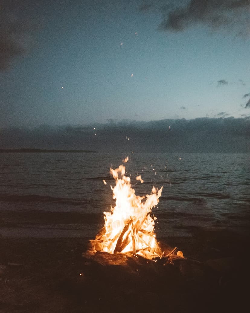 Burning campfire on a sandy beach with crashing ocean waves under a dark blue twilight sky at the shoreline.