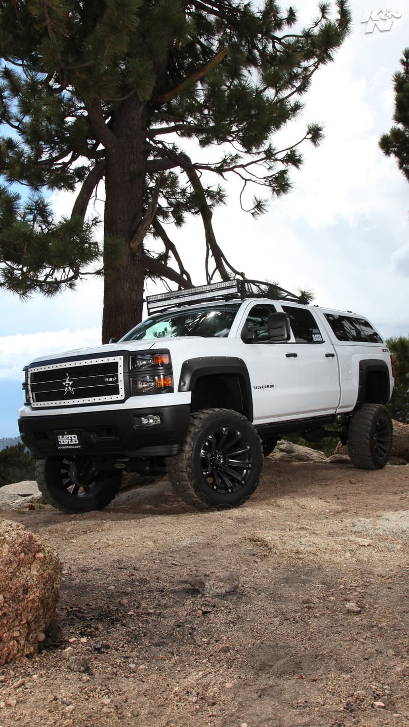 Lifted white Chevrolet Silverado with black off-road wheels and a light bar parked on a rocky mountain slope.