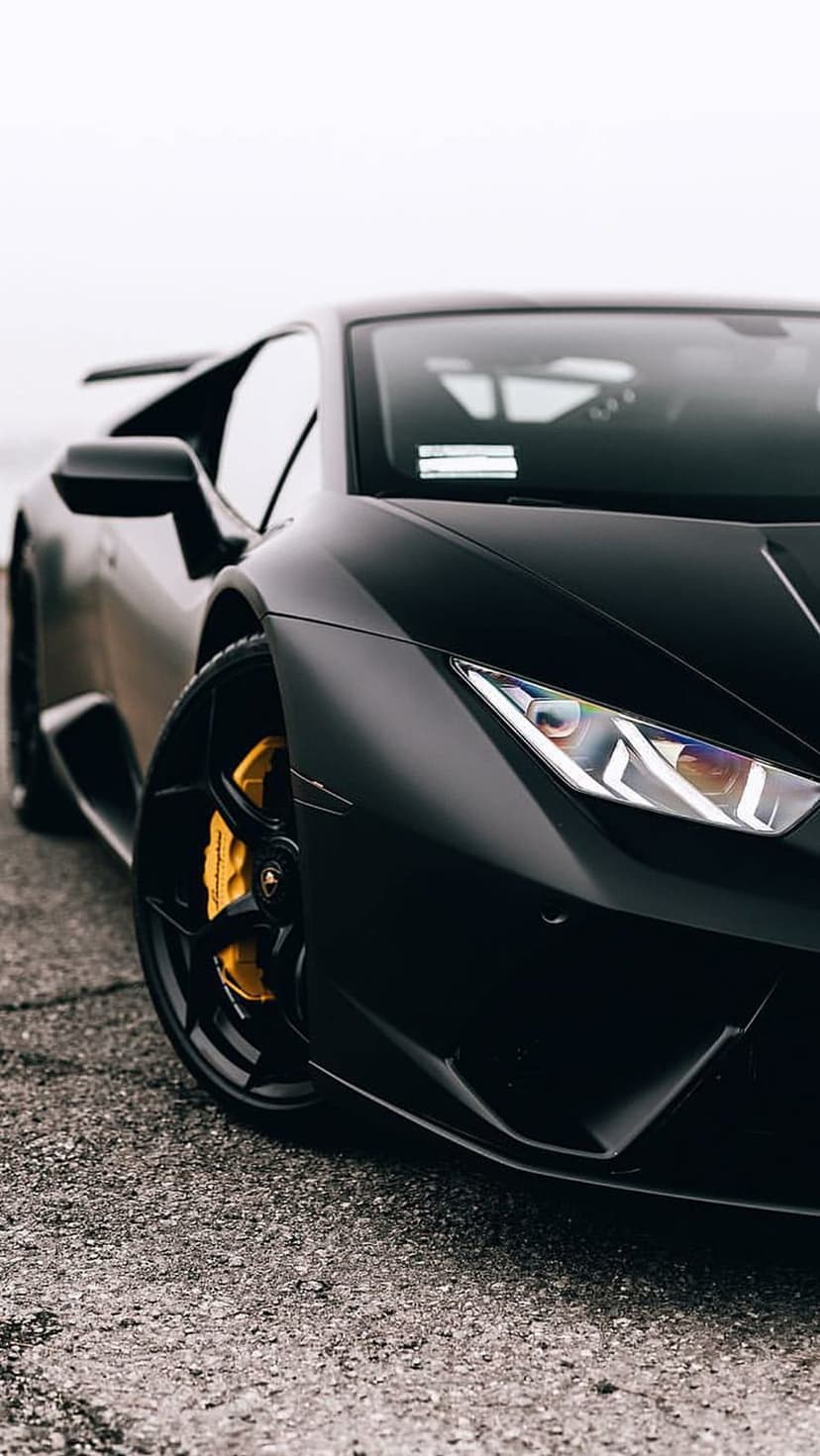 Matte black Lamborghini Huracan with glowing LED headlights and yellow brake calipers on dark textured asphalt.