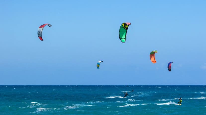 Kite surfers on boards ride choppy turquoise waves under a sky filled with red, green, and orange kites.