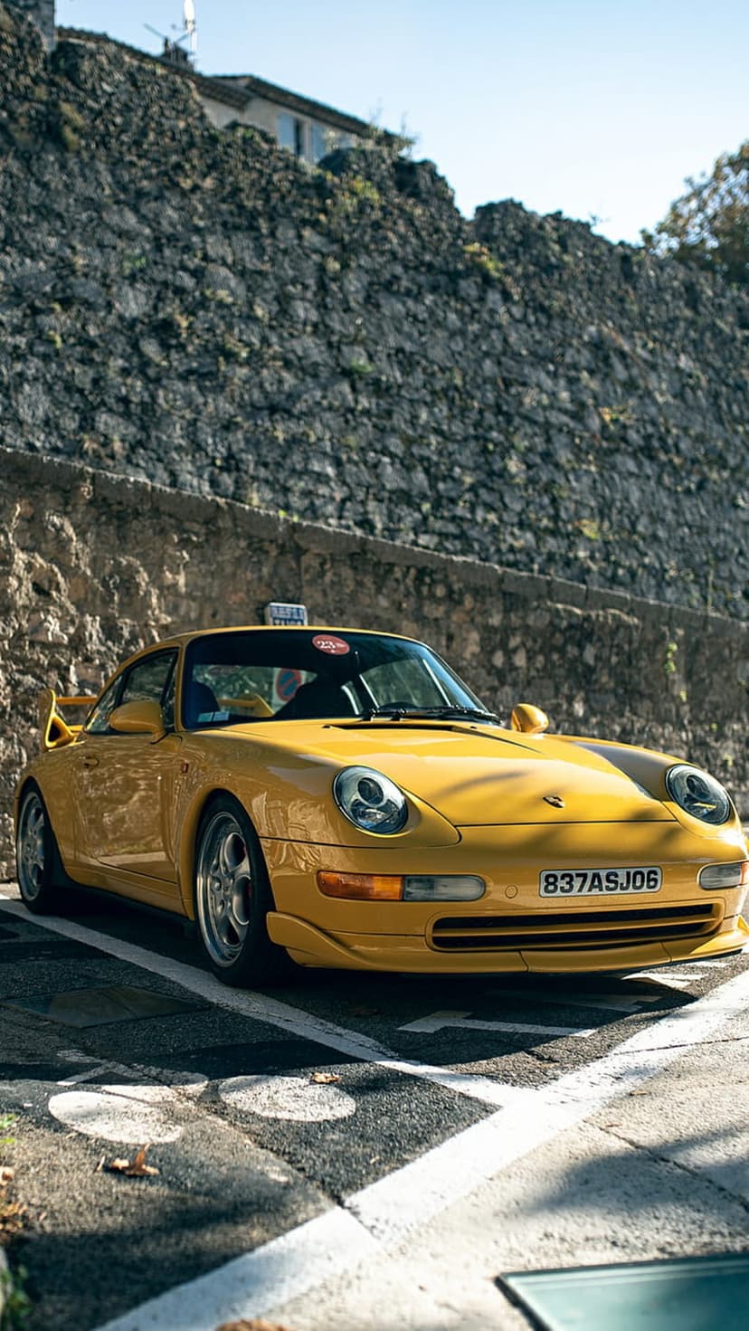 Yellow Porsche 911 with a taxi sign on its roof parked along a textured stone wall in bright sunlight.